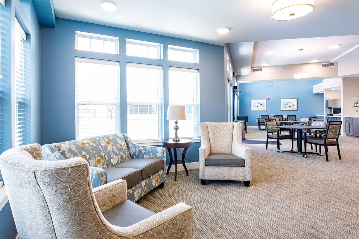 A cozy living room with modern furniture, featuring a sofa, chairs, and a table set against blue walls and large windows.