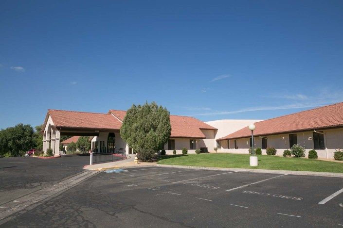 A single-story building with a red roof set against a clear blue sky, featuring a parking lot and landscaped area with a tree.