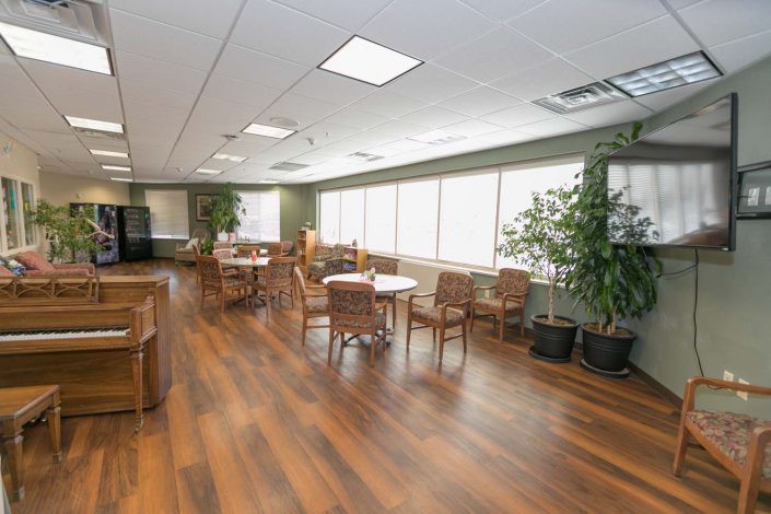A spacious lounge area with tables and chairs, featuring a wooden floor and large windows allowing natural light to enter.