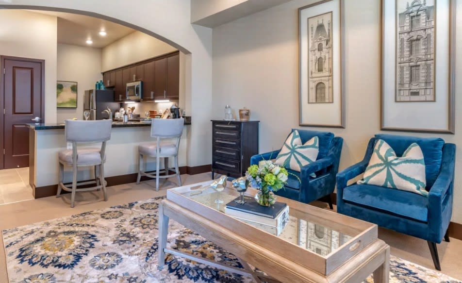 A modern living room with a cozy atmosphere, featuring blue chairs, a decorative coffee table, and an open view into the kitchen.