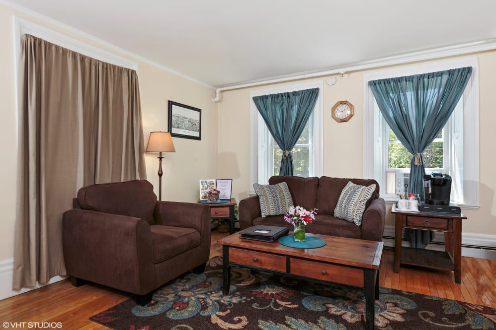 A cozy living room with a brown couch and armchair, a wooden coffee table, and colorful curtains.