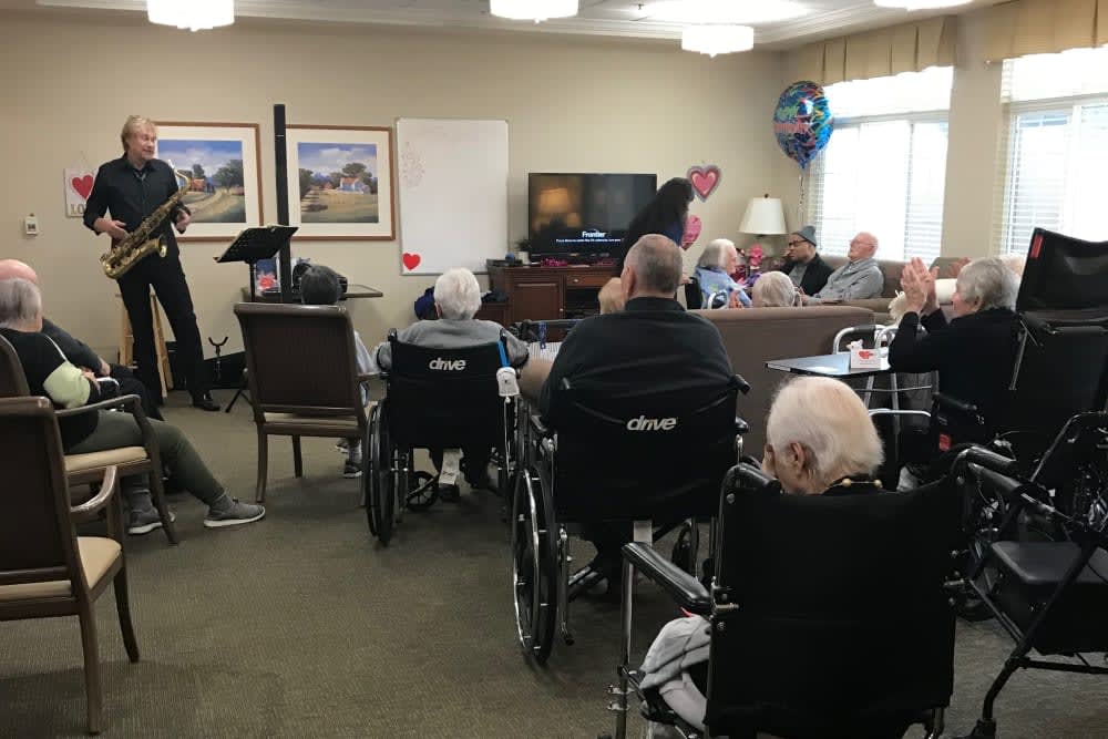 A group of elderly people gathered in a nursing home for a communal activity, some seated in wheelchairs.