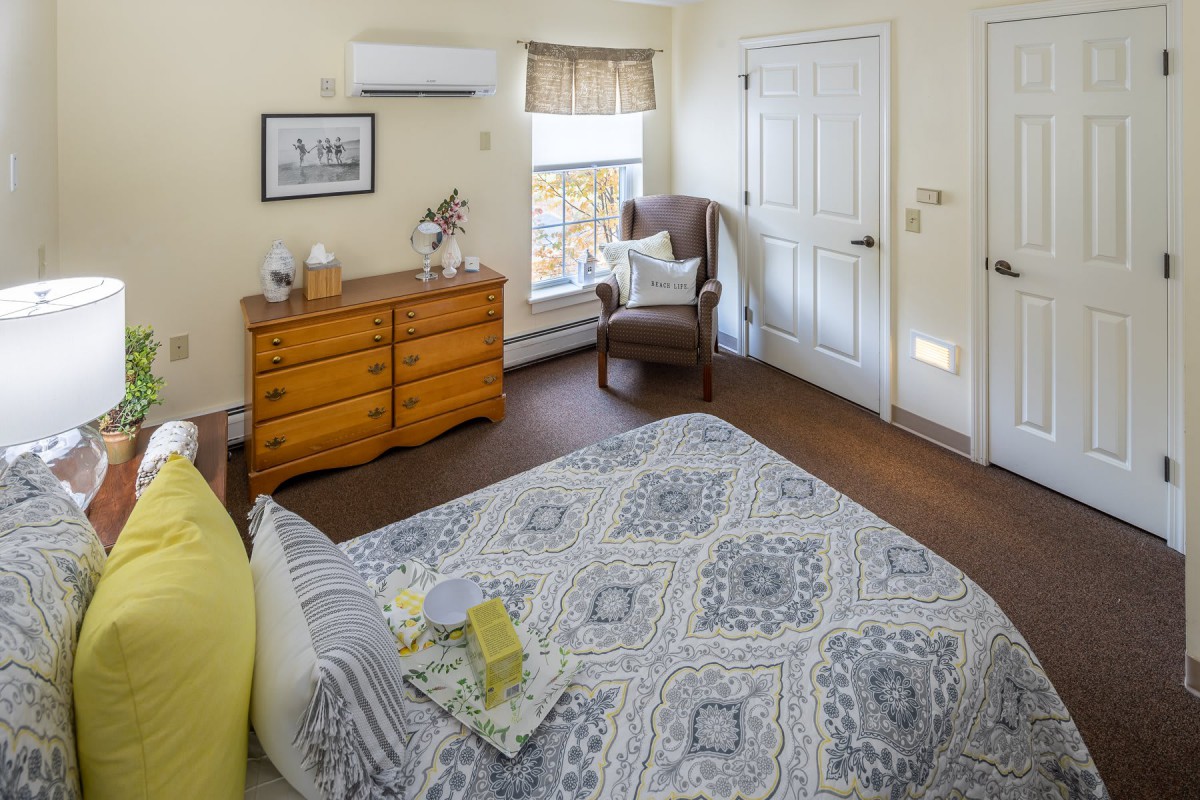 A cozy bedroom with a patterned bedspread, wooden dresser, and armchair by the window.