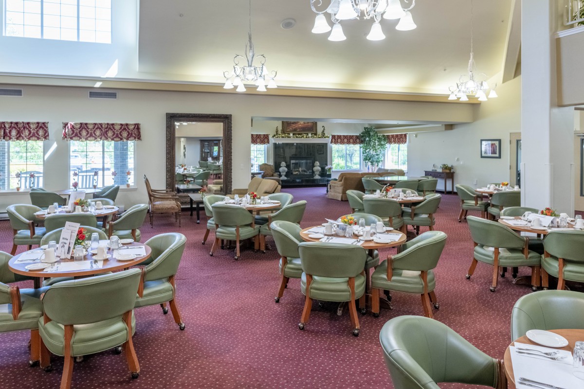 A spacious dining room with round tables set for a meal, featuring elegant chandeliers and green chairs on a red carpet.