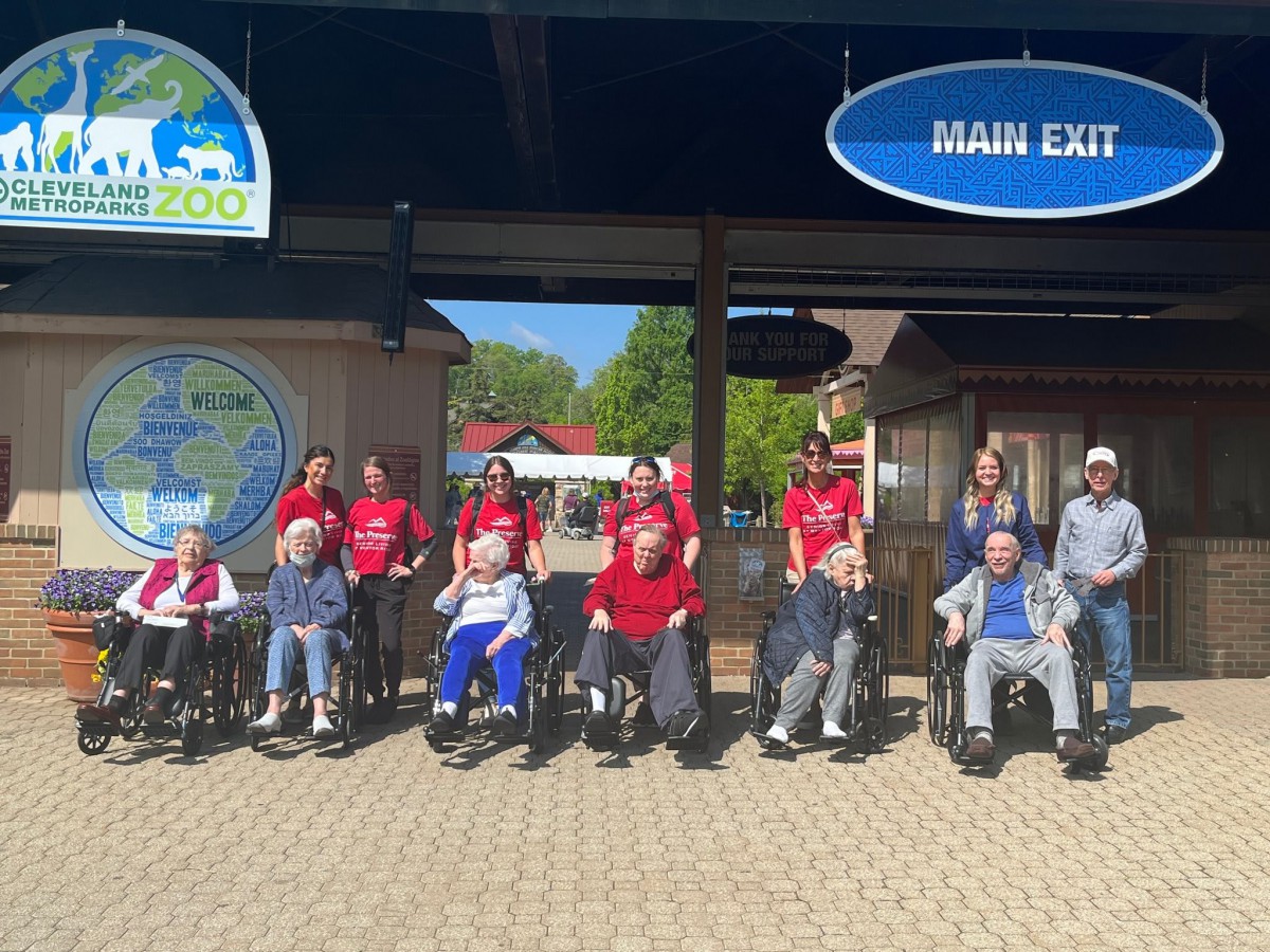 A group of people in wheelchairs with others standing behind them, gathered at an outdoor entrance.