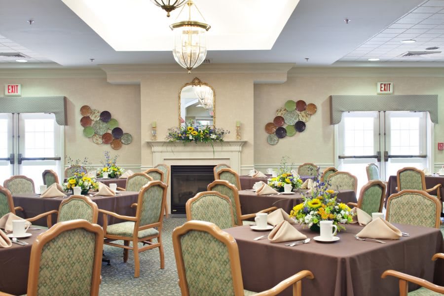 A well-decorated dining room with tables set for a meal, featuring elegant chairs, floral centerpieces, and a fireplace.
