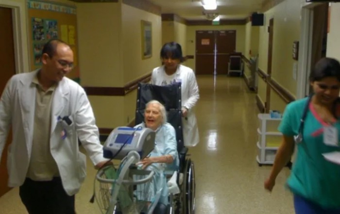 Healthcare workers assist an elderly woman in a wheelchair down a hospital corridor.