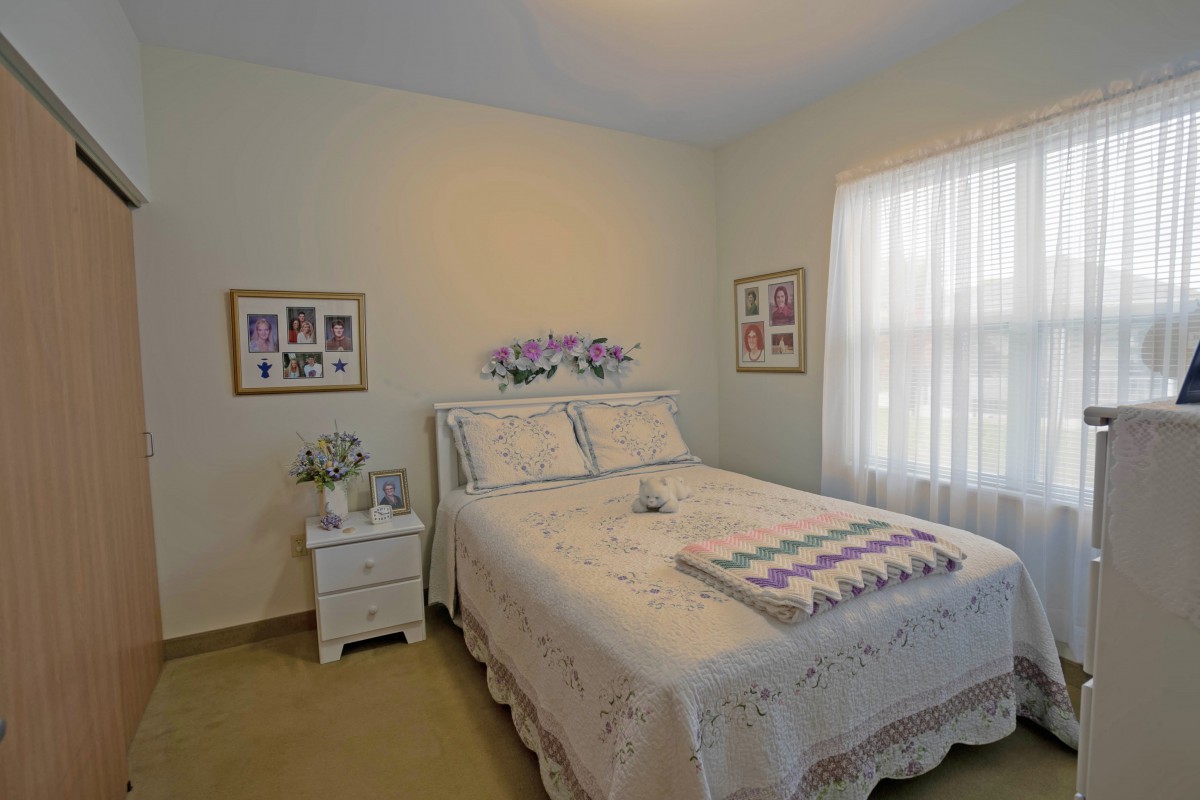 A cozy bedroom with a neatly made bed adorned with floral bedding and decorative pillows, accompanied by framed pictures on the wall and a sunlit window.
