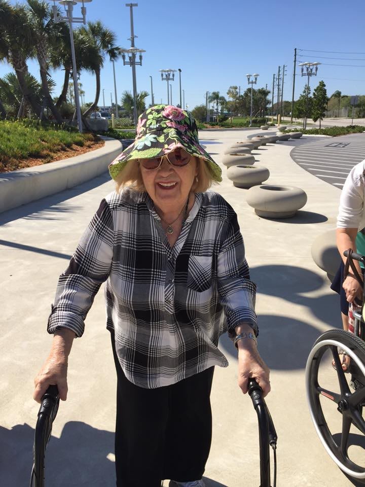 An elderly person enjoys a sunny day outdoors, using a walker and wearing a floral hat and plaid shirt.