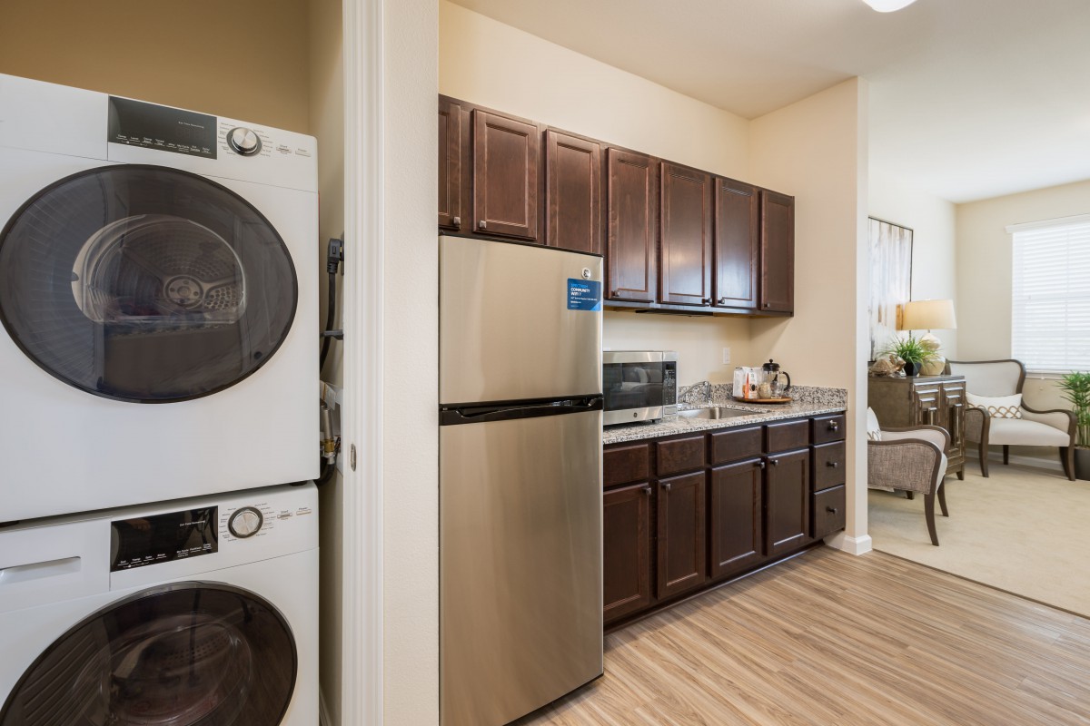 A modern kitchen with stacked washer and dryer, stainless steel refrigerator, microwave, and dark wood cabinets.
