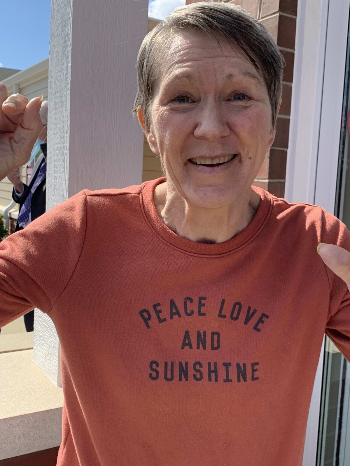 A person smiling while wearing a shirt with the message 'Peace, Love, and Sunshine' outdoors.