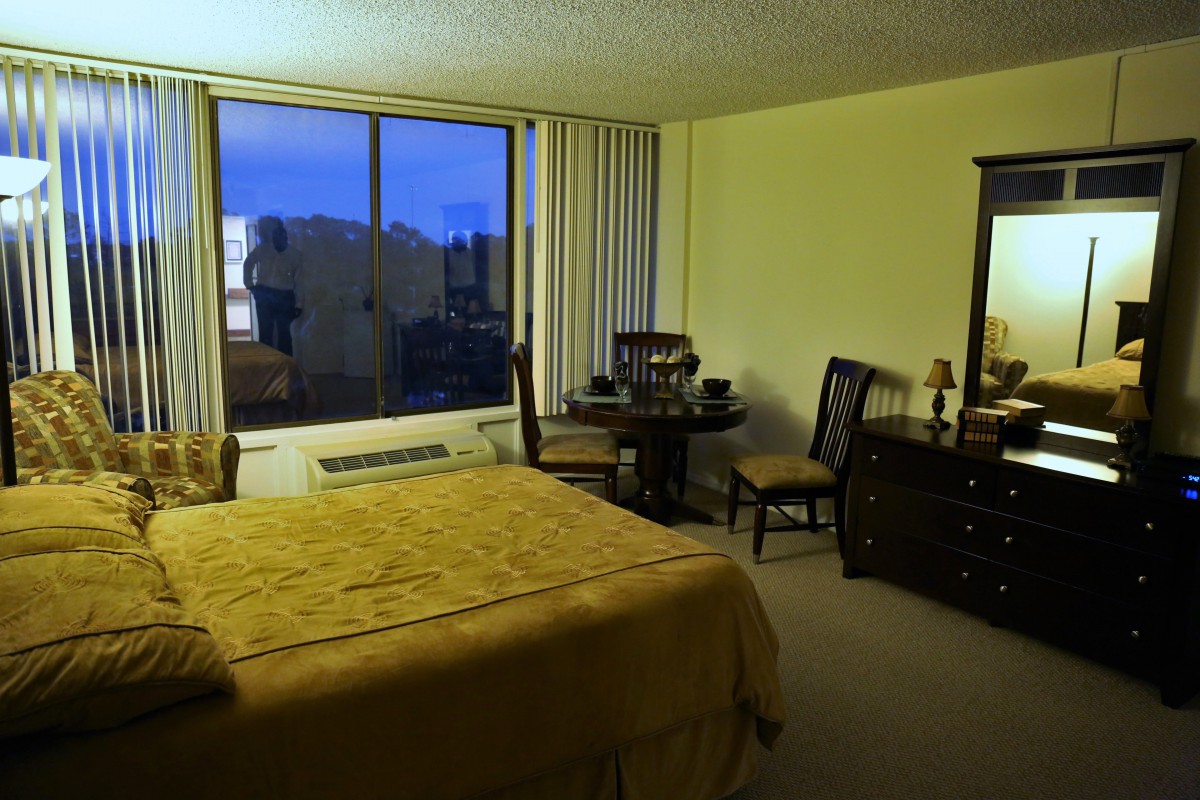 A cozy hotel room with a large bed, wooden furniture, and a window view during the evening.