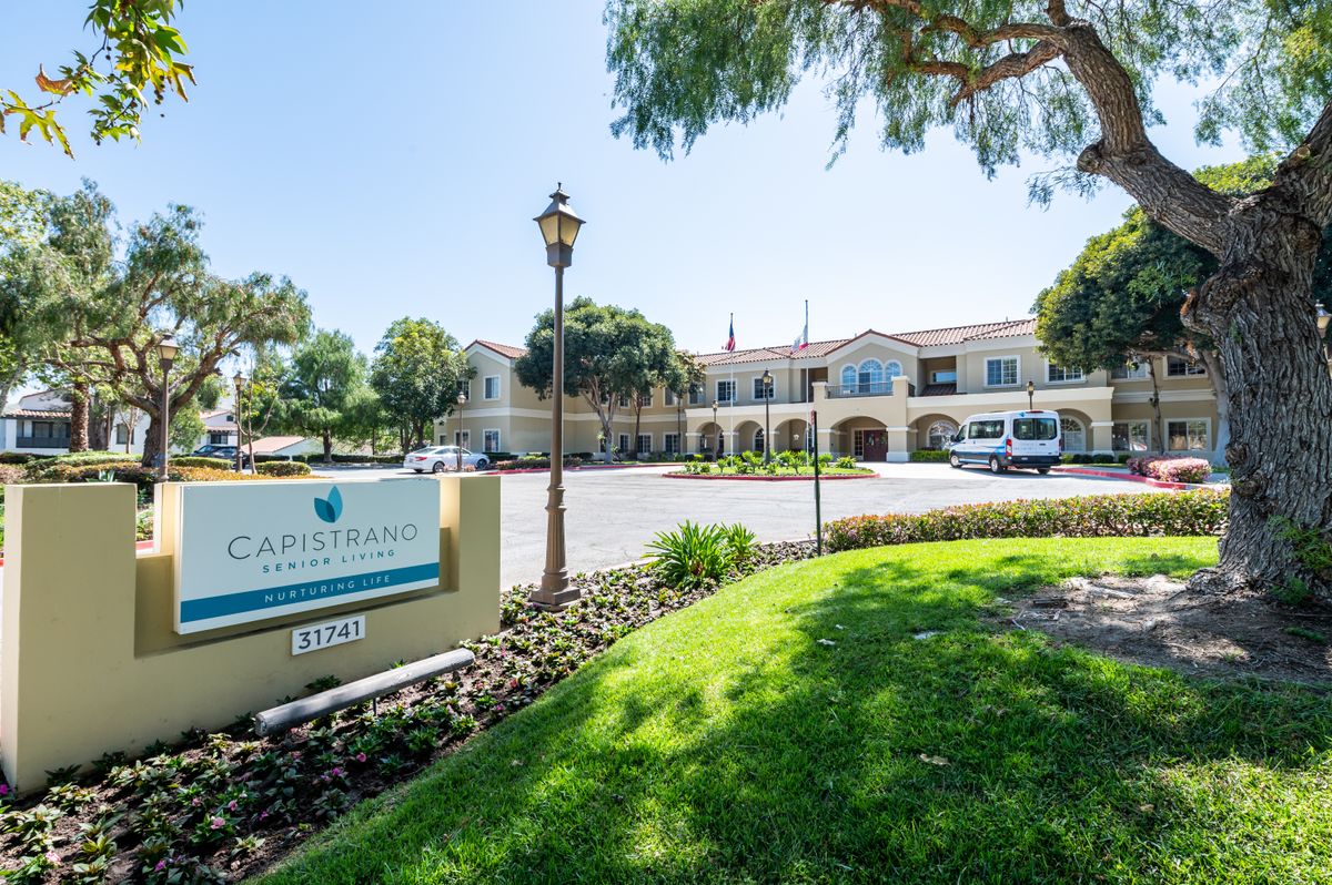 A sunny view of a large building with a sign on a landscaped lawn and a parking lot in front.