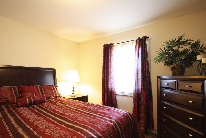 A cozy bedroom with a striped bedspread, lamp, and dark curtains filtering sunlight through the window.