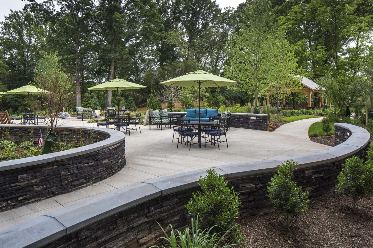 A landscaped outdoor patio area with tables, chairs, and green umbrellas surrounded by a stone wall and greenery.