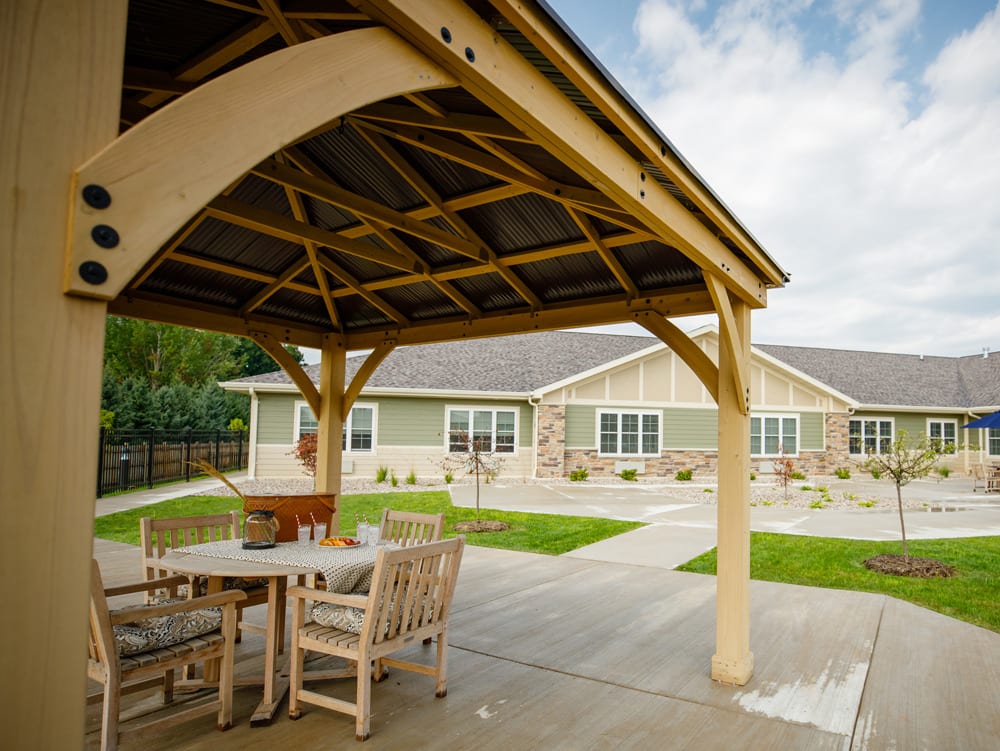 A cozy outdoor gazebo with wooden furniture on a patio, set against the backdrop of a modern single-story building.
