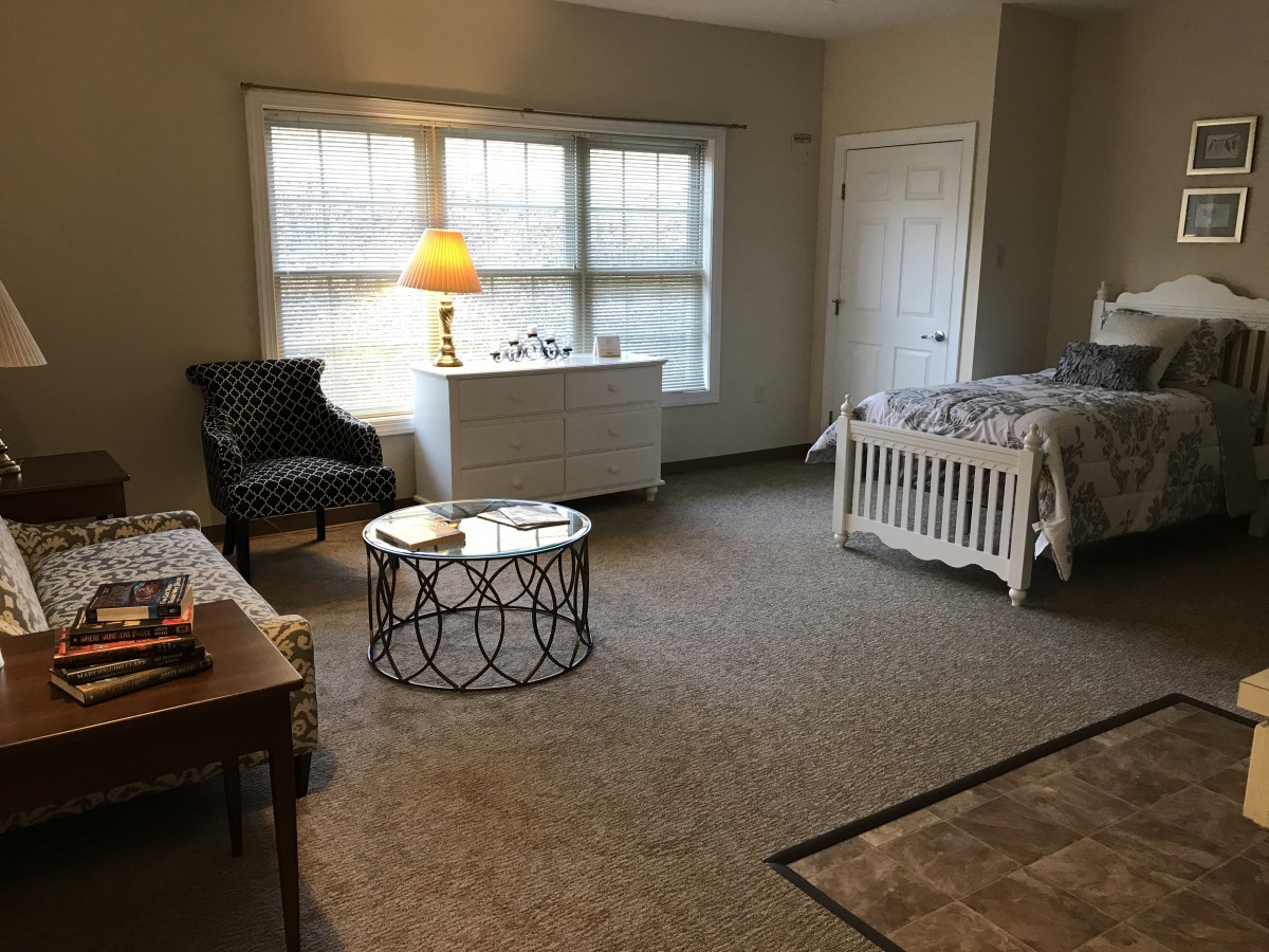 A cozy bedroom featuring a bed, chair, and side table with a lamp near a window.