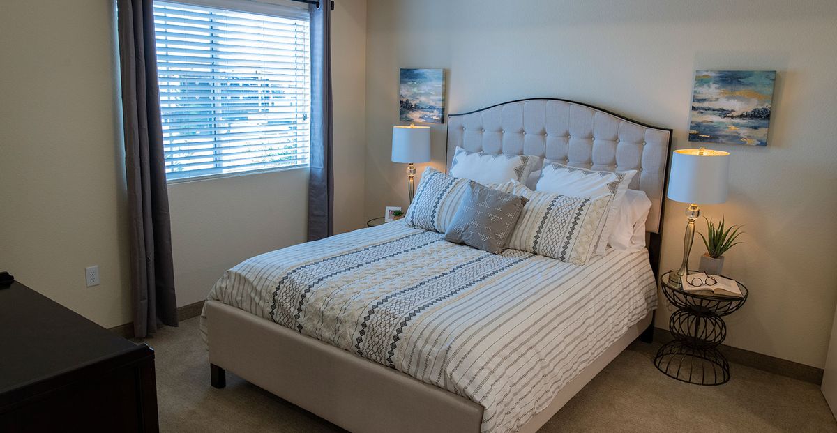 A cozy bedroom with a neatly made bed featuring striped bedding, a bedside lamp, and a window with blinds and curtains.