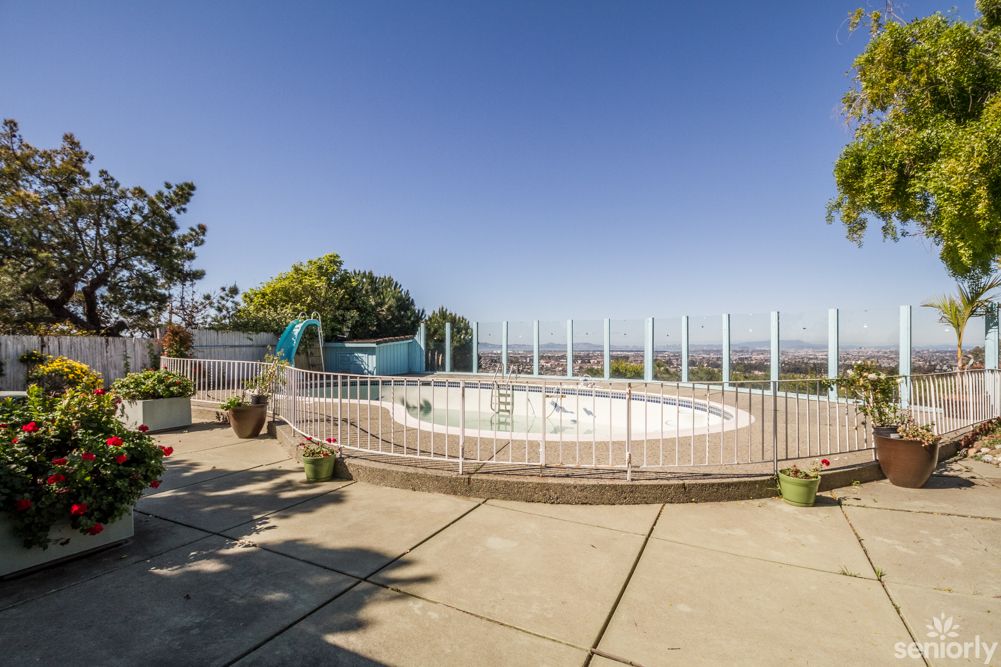 An empty, fenced outdoor pool surrounded by a patio and potted plants under a clear blue sky.