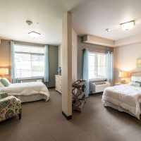 A well-lit bedroom featuring two beds, windows with curtains, and carpeted flooring.