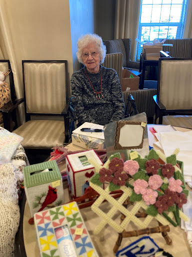An elderly woman seated at a table surrounded by crafts and decorative items.