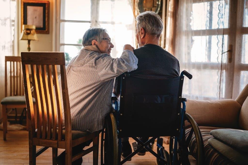Two elderly individuals sitting together, one in a wheelchair, sharing a moment of companionship by the window.