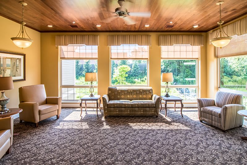 A cozy living room with ample natural light streaming through large windows, featuring comfortable seating and a detailed carpet.