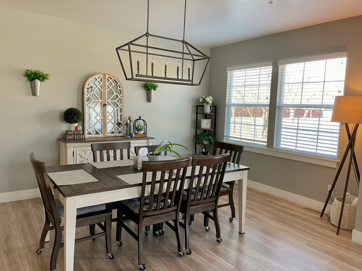 A cozy dining room with a wooden table and chairs, illuminated by a decorative chandelier, with natural light streaming in through the windows.