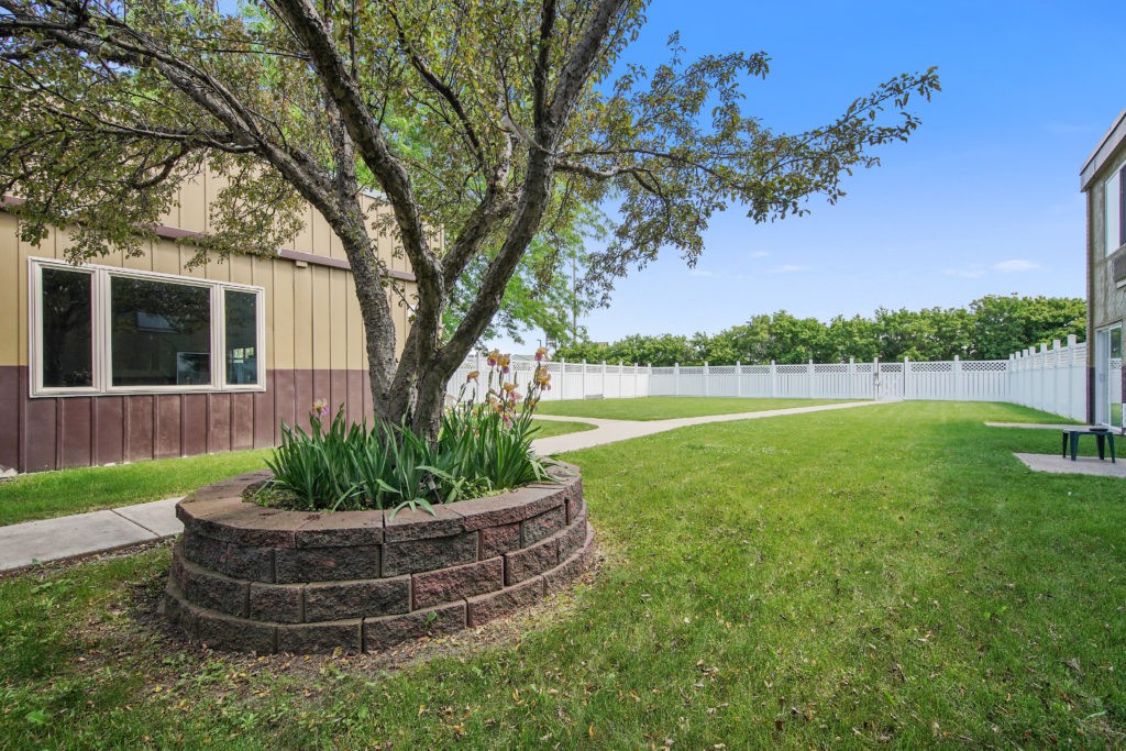 A well-maintained lawn with a tree surrounded by a circular brick planter, adjacent to a building and white fence.