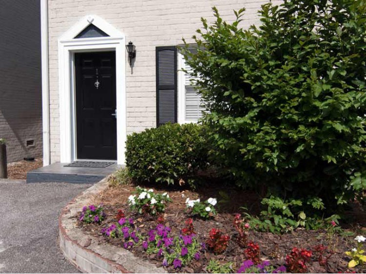 A house entrance with a black door, surrounded by a well-maintained garden featuring vibrant flowers and green bushes.