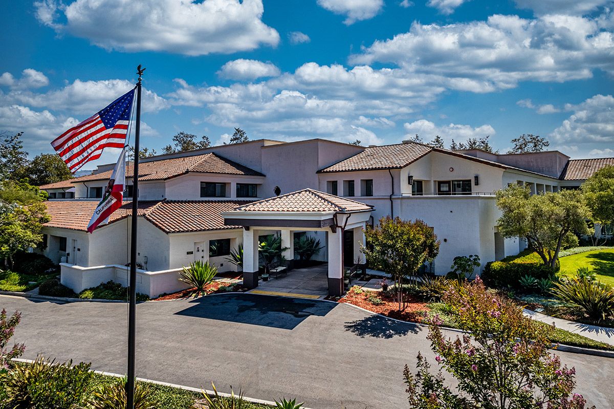 A large residential house with a driveway, surrounded by landscaped gardens and an American flag in the foreground under a partly cloudy sky.
