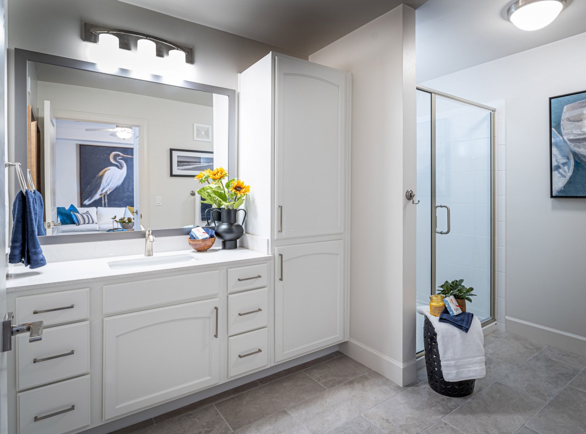 A modern bathroom with white cabinets, a large mirror, and a glass shower enclosure.