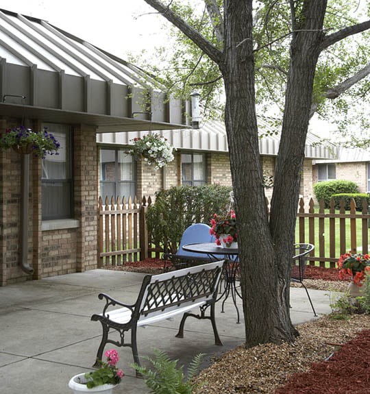 A peaceful courtyard features a bench and table set near a tree with surrounding fences.
