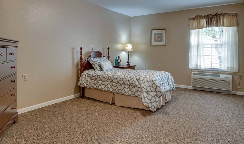 A well-lit bedroom with a single bed, nightstand, and a lamp by the window.
