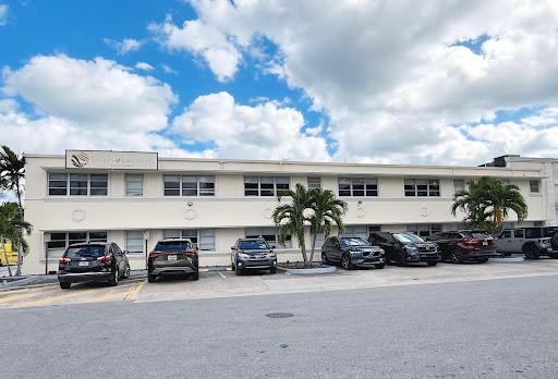 A view of a multi-story building with parked cars in front and palm trees under a cloudy sky.