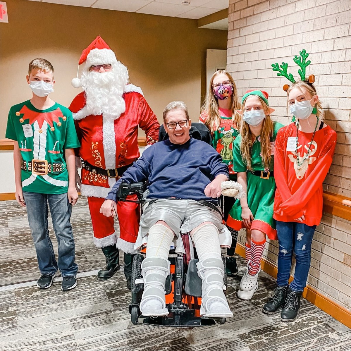 A group of people celebrating the holidays with festive outfits, including Santa Claus, gathered around a person in a wheelchair.