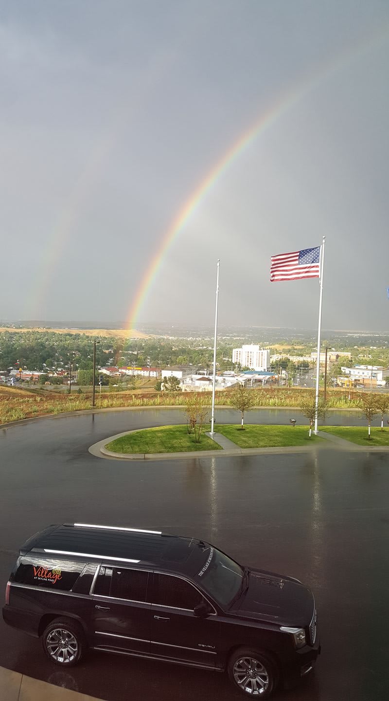 A double rainbow arches over a landscape with an American flag waving in the foreground.