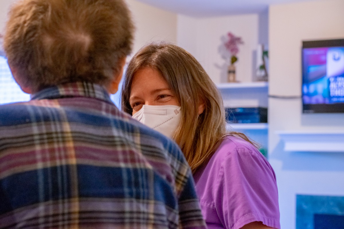 A healthcare professional wearing a mask engages in conversation with an elderly person.