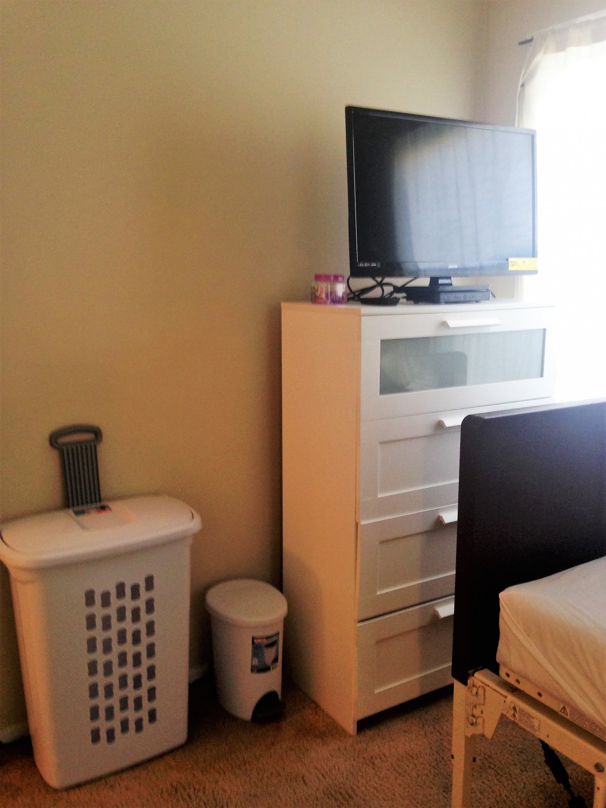 A bedroom corner featuring a dresser with a TV on top, accompanied by a laundry basket and trash can.