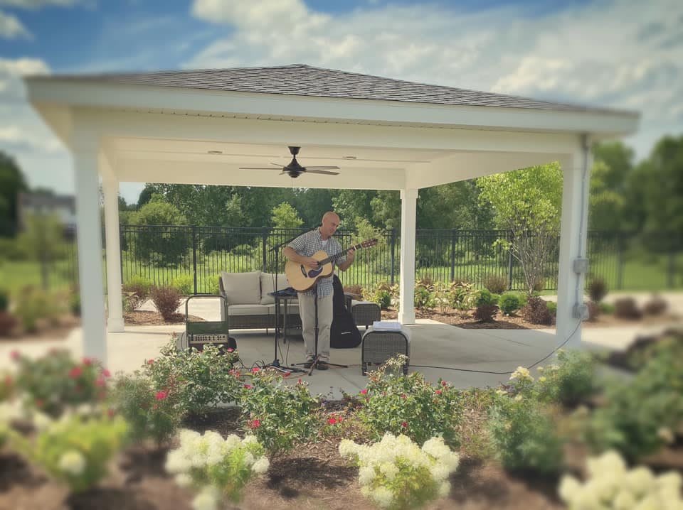 A musician playing guitar in a gazebo surrounded by a garden.