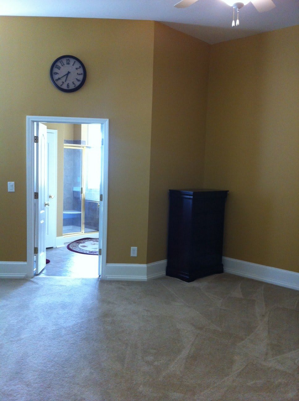 An empty room with beige walls, carpet flooring, a clock on the wall, and a doorway leading to another area.