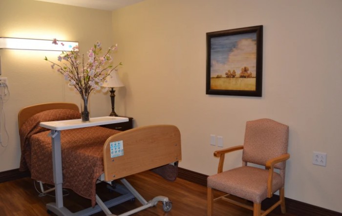 A cozy hospital room with a bed, chair, and decorative items including a painting and flowers.