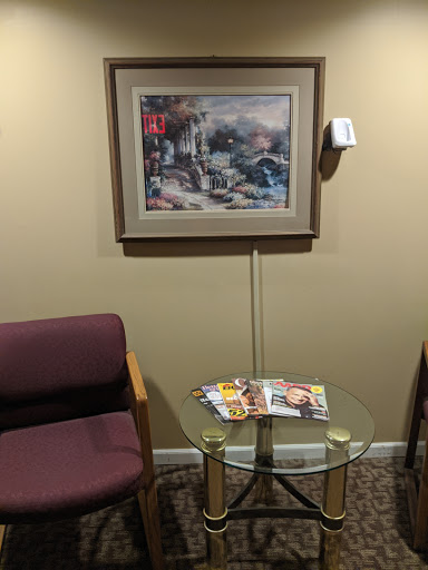 A cozy waiting area featuring a framed picture on the wall, a glass table with magazines, and two chairs.