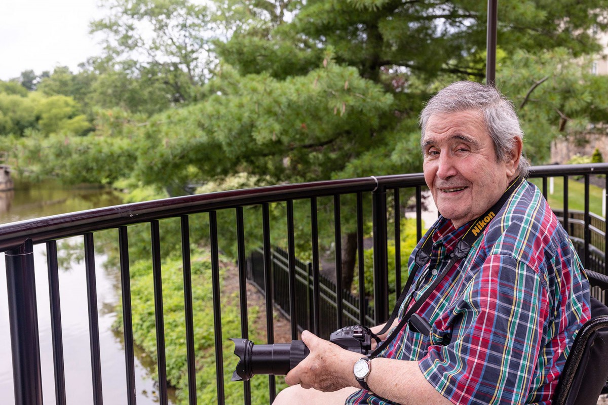An elderly man in a wheelchair, smiling while holding a camera, enjoying the view of nature from a balcony.