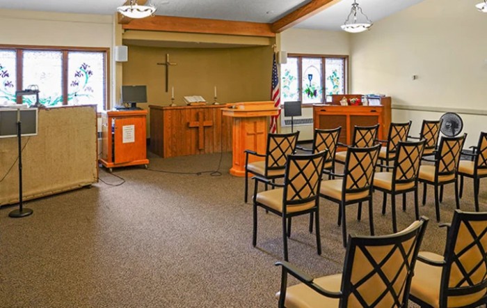 An empty chapel interior with rows of chairs facing a pulpit and cross.