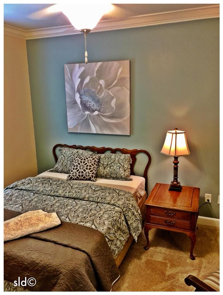A cozy bedroom featuring a neatly made bed, a wooden nightstand with a lamp, and a large abstract painting on the wall.