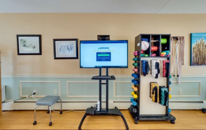 A physical therapy room featuring a monitor on a stand and a variety of exercise equipment neatly organized.