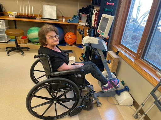An elderly woman engaging in rehabilitation exercises using a specialized stationary bike while seated in her wheelchair.