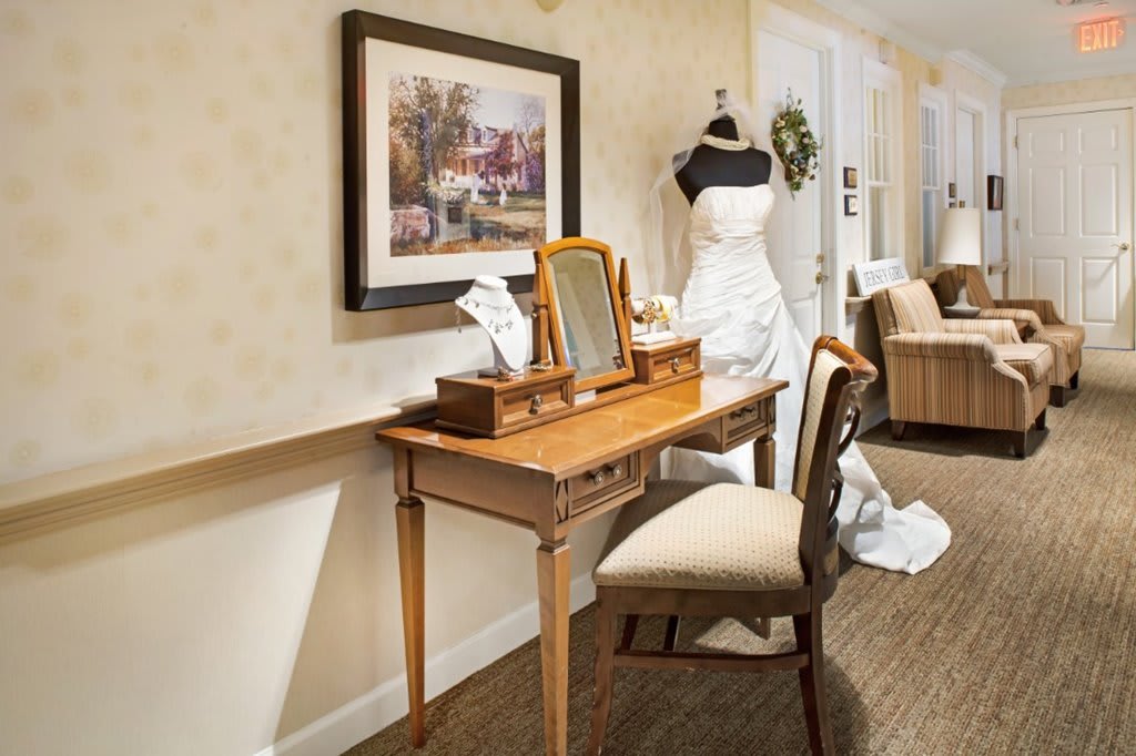 A vintage-style vanity setup featuring a mirror and chair, with a wedding dress on a mannequin in the background.