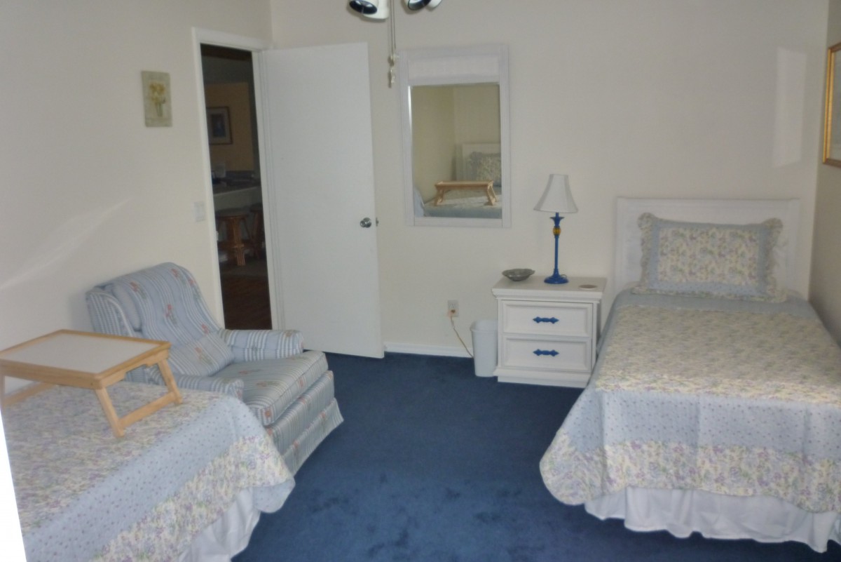 A cozy bedroom featuring twin beds, a chair, and a nightstand with a lamp on a blue carpet.